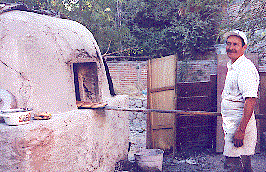 [Baker taking bread from large outside mud brick oven in the backyard of his bakery/home: 27k]