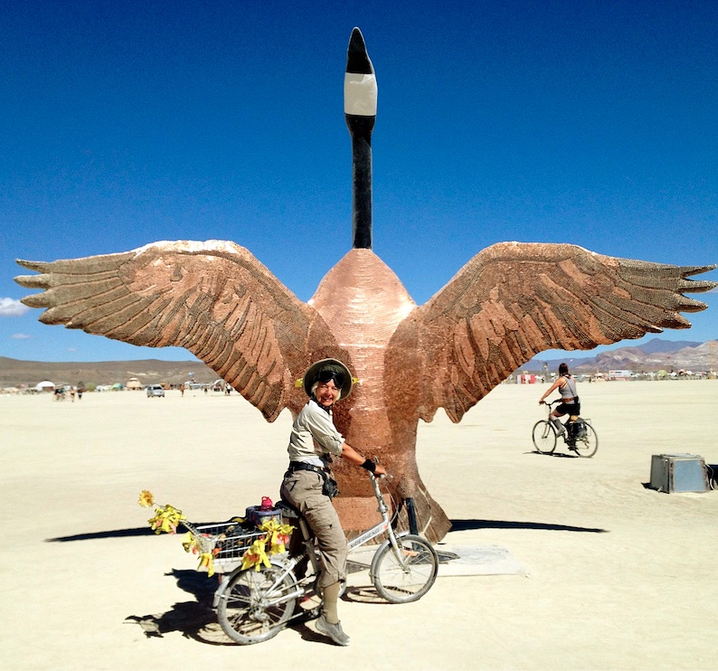 [Carolyn on bicycle next to towering Canadian goose made from copper pennies at Burning Man 2015: 78k]