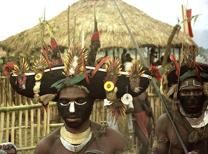 [Two wigmen wearing dramatic wide black hair wigs decorated with multi-colored brilliant Bird of Paradise plumes, including a complete bird, black face paint and kina shells, stand before their thatched ramada and bamboo fence: 340k]